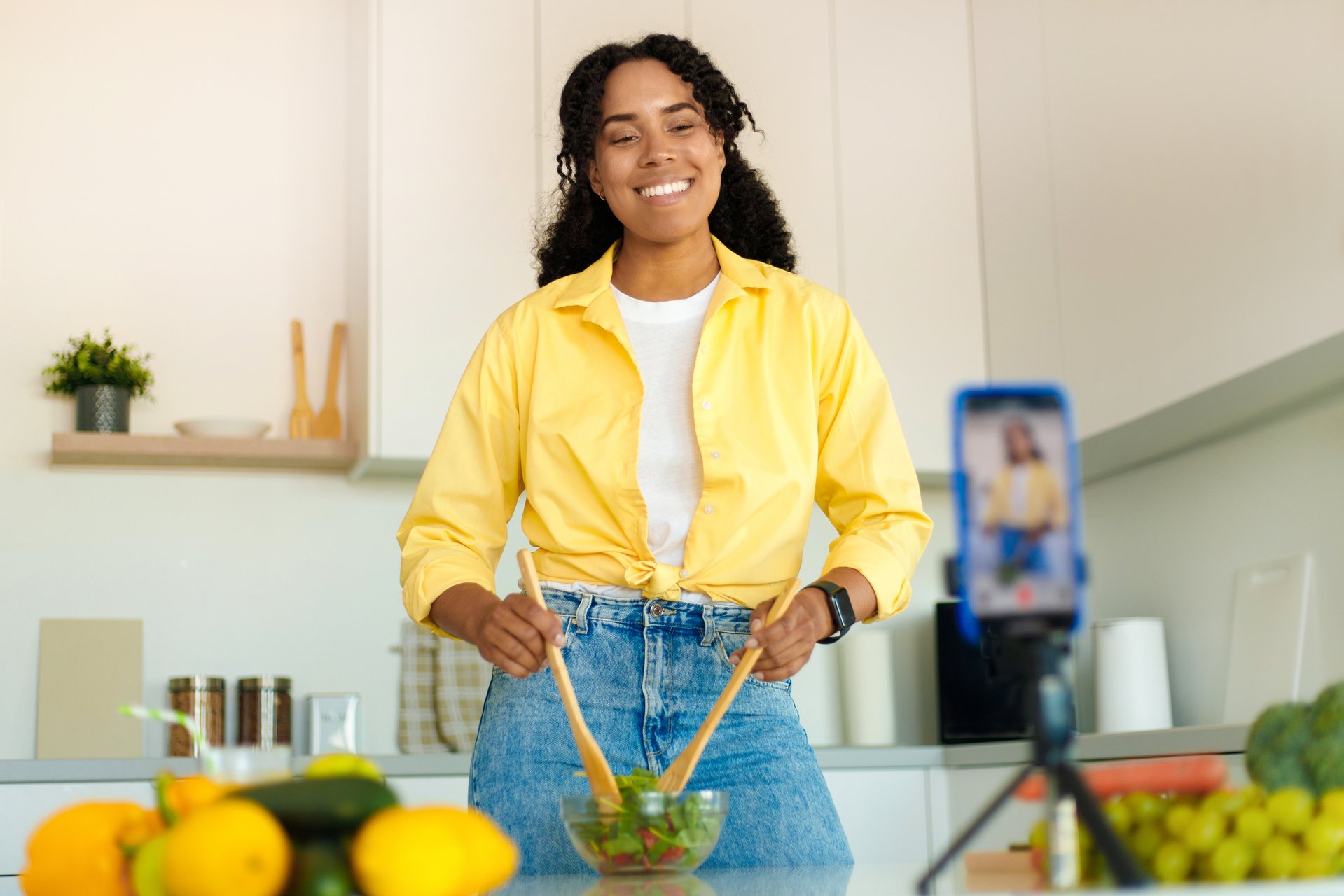 Nutrition coach. Excited black woman recording video recipe in kitchen, using smartphone on tripod